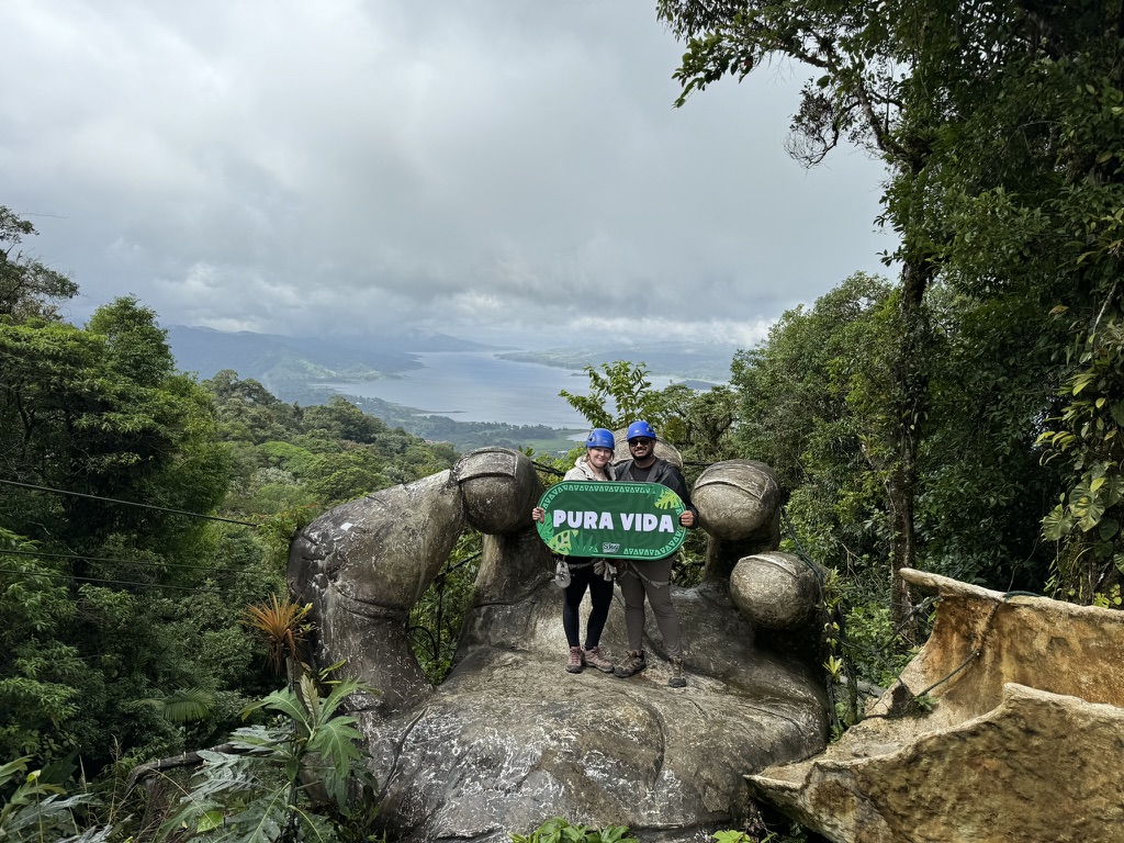 Surya and Leslie ziplining in Costa Rica, holding a Pura Vida sign above a rainforest lake
