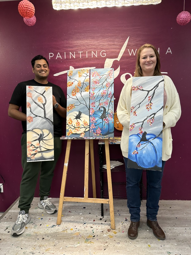 Surya and Leslie holding their autumn-themed canvases at a paint-and-sip class